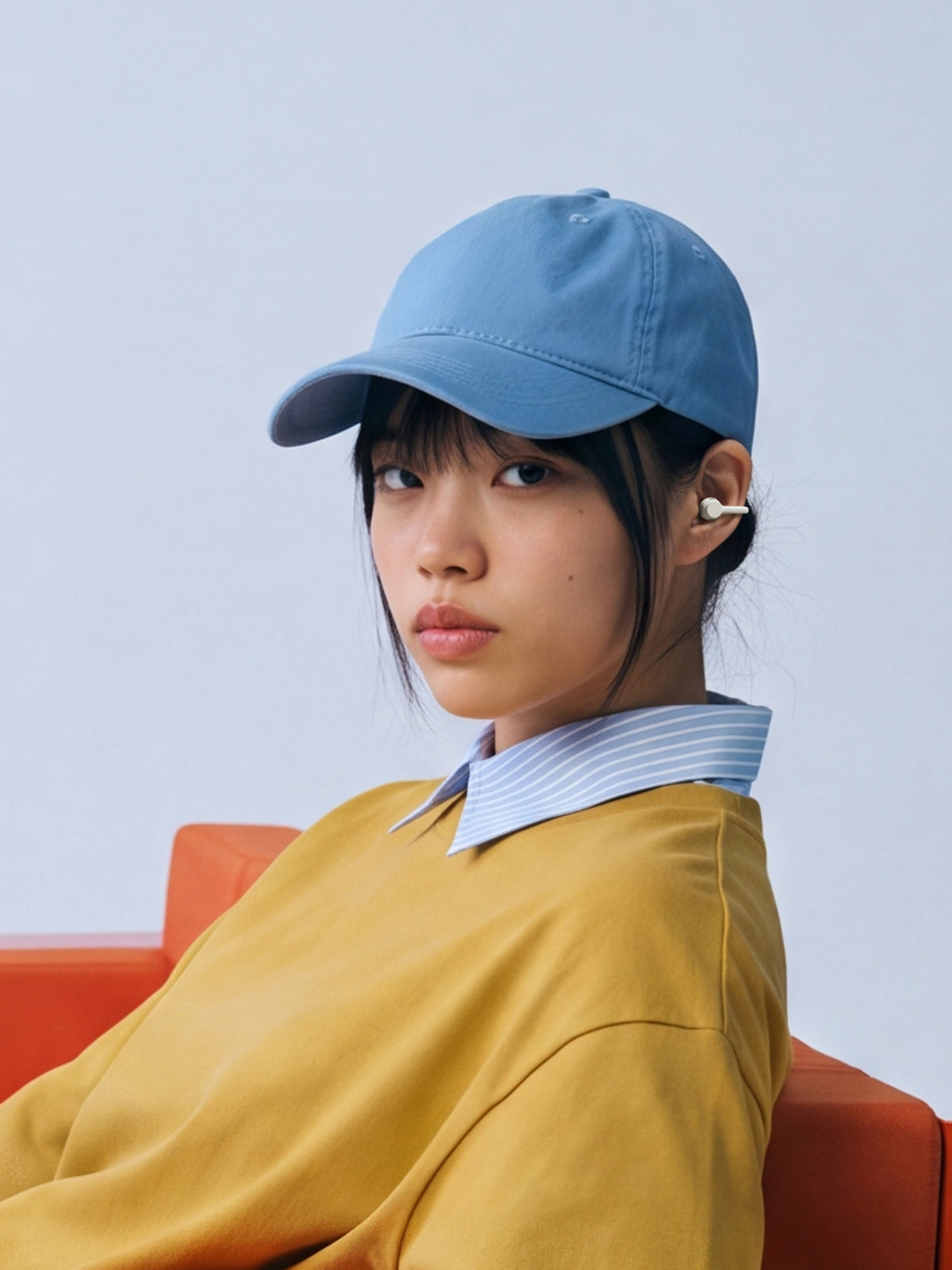 A woman in a cap and yellow top seated on an orange chair wearing a clip earbud