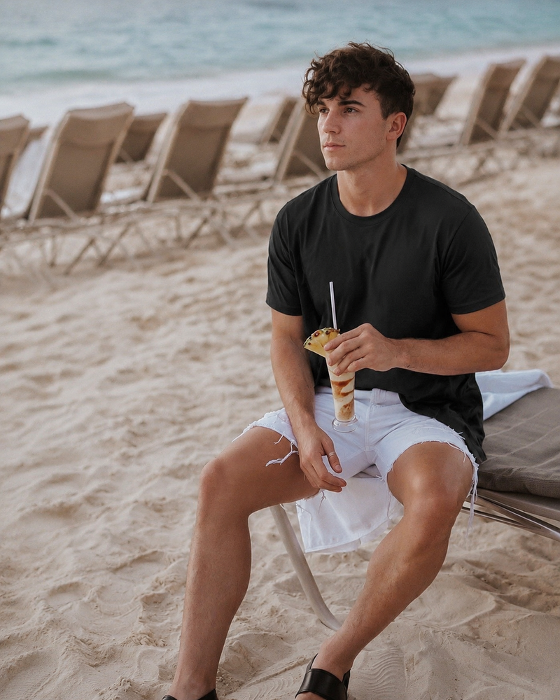 Model in a black relaxed tee and white shorts, seated beside the beach with a drink.