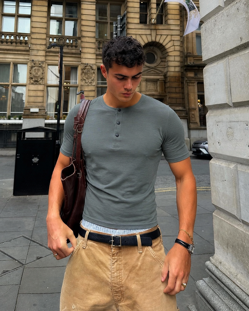 Model wearing a sage green snap-neck Henley with tan trousers on a city street.
