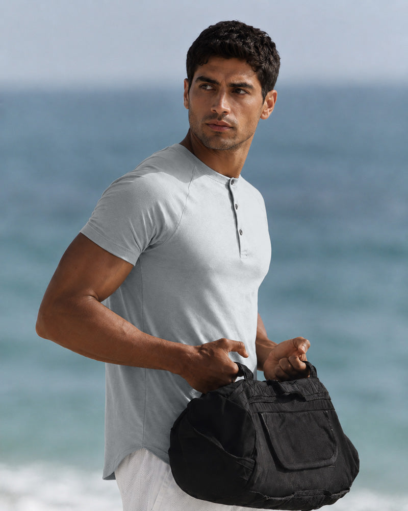 Model wearing the Overcast light grey Henley beside the sea, carrying a black duffle bag.